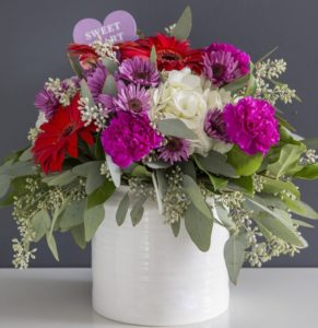 This white ceramic contains white hydrangea, assorted holiday gerberas, assorted holiday carnations, assorted holiday poms, assorted eucalyptus, and salal 